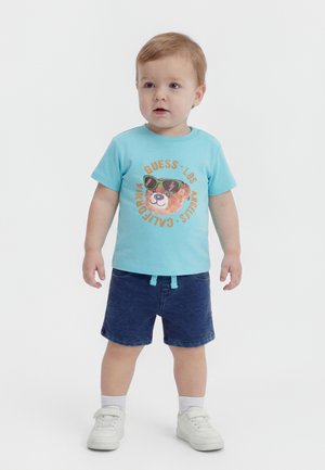 Νήπιο που στέκεται φορώντας μπλε μπλουζάκι με σχέδιο αρκούδας, σκούρα μπλε σορτς, λευκές κάλτσες και λευκά αθλητικά παπούτσια, σε λευκό φόντο.