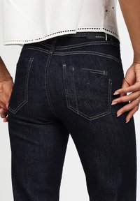 Vaqueros de mezclilla azul oscuro con un corte ajustado. Presentan dos bolsillos traseros, costuras en contraste y un parche de logo de marca negro en la cinturilla.