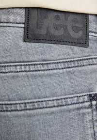 Tela de mezclilla gris con costuras visibles, que presenta una etiqueta de cuero negro con texto en relieve cosida en la cinturilla en la parte posterior.