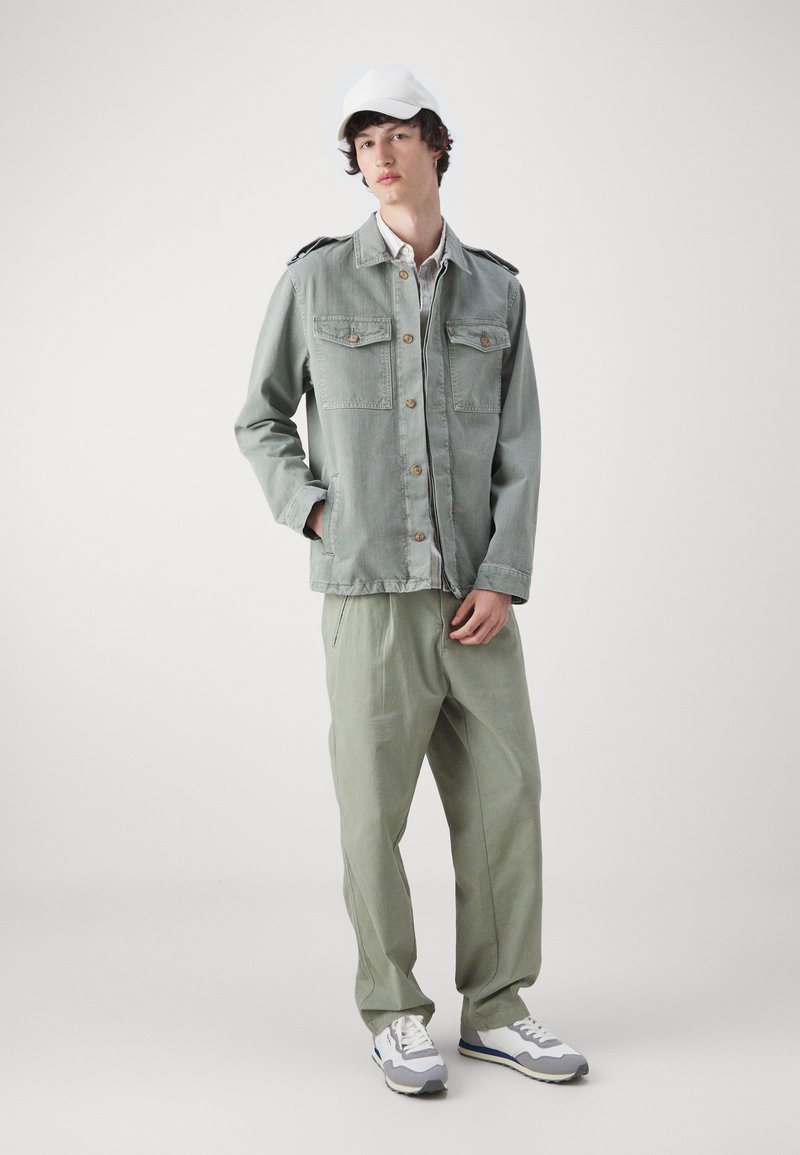 Chaqueta de mezclilla gris claro con dos bolsillos en el pecho y cierre de botones, combinada con pantalones sueltos de color verde claro y zapatillas blancas.