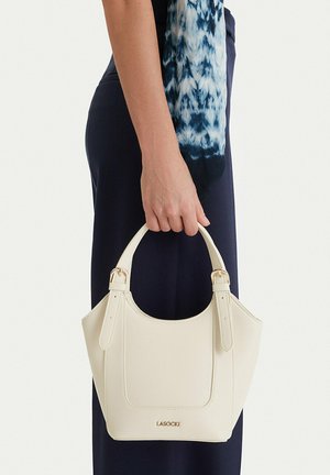 Persona sostiene un pequeño bolso blanco Lasocki por las asas superiores contra un fondo de pantalones oscuros y una blusa azul con estampado.