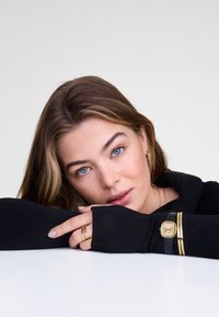 Haut noir à manches longues avec texture côtelée, présentant un col plié. Bracelet en cuir noir avec des accents dorés, bagues empilées sur les doigts.