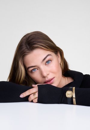 Haut noir à manches longues avec texture côtelée, présentant un col plié. Bracelet en cuir noir avec des accents dorés, bagues empilées sur les doigts.