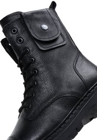 Bota de combate de cuero negra con un pequeño bolsillo con solapa, cordones negros redondos, ojales y una suela gruesa con dibujo.