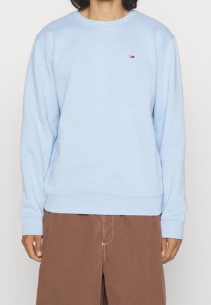 Personne portant un sweat-shirt bleu clair à col rond avec un petit logo Tommy Hilfiger sur la poitrine et un pantalon marron.