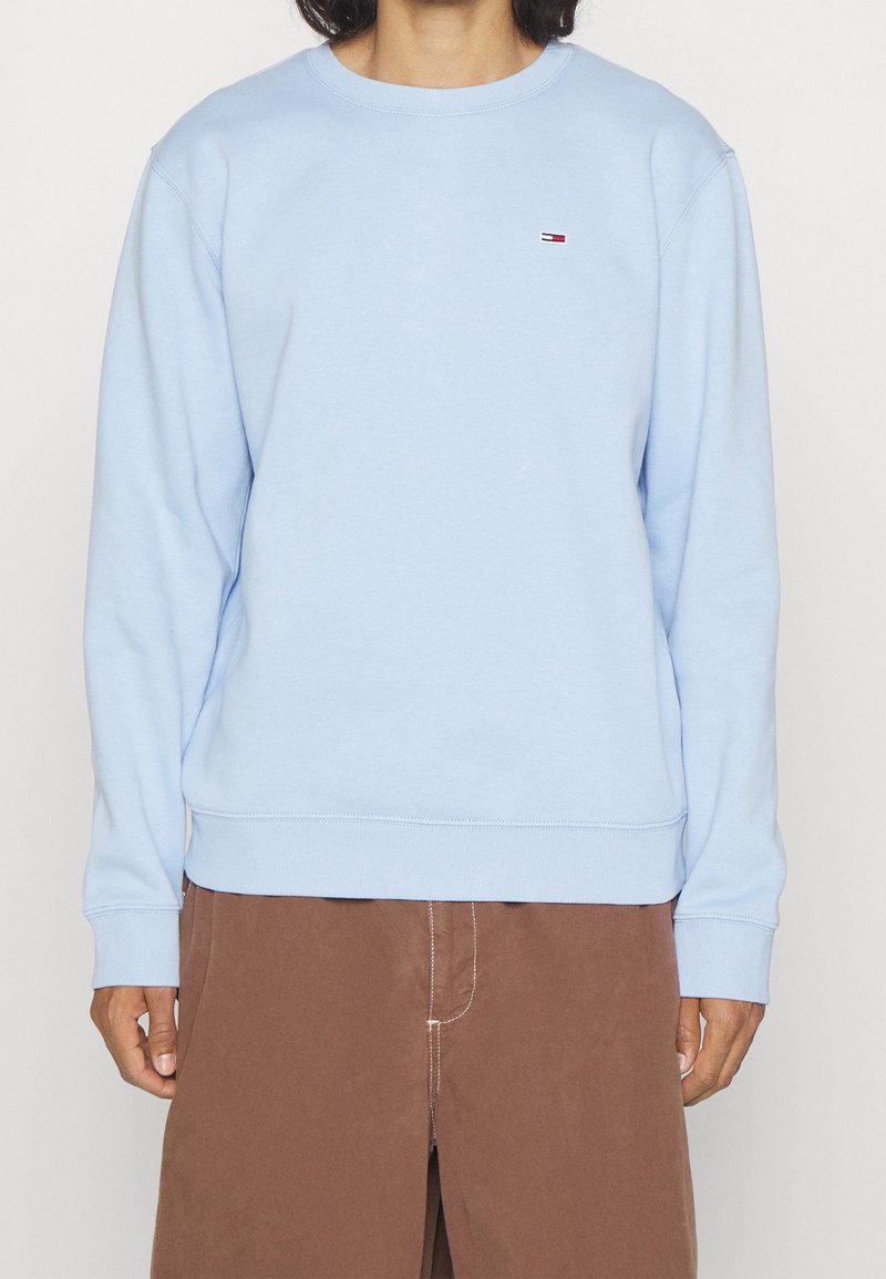 Personne portant un sweat-shirt bleu clair à col rond avec un petit logo Tommy Hilfiger sur la poitrine et un pantalon marron.