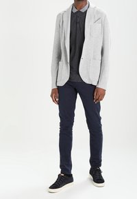 Grauer Jersey-Blazer mit zwei Fronttaschen über einem dunkel gepunkteten Hemd, kombiniert mit marineblauen Slim-Fit-Hosen und schwarzen Turnschuhen.