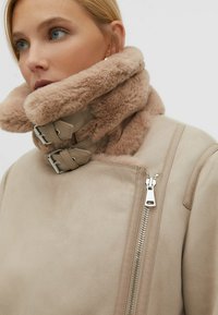 Beige Jacke mit Kunstfellkragen verfügt über ein glattes Wildlederfinish, zwei Schnallen und einen seitlichen Reißverschluss. Der Kragen ist plüschig und übergroß.