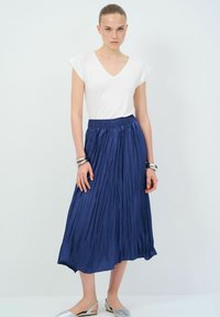 Jupe midi plissée bleu marine avec taille élastique, assortie à un haut ajusté blanc à col en V et des ballerines argentées. Texture lisse, design minimaliste.