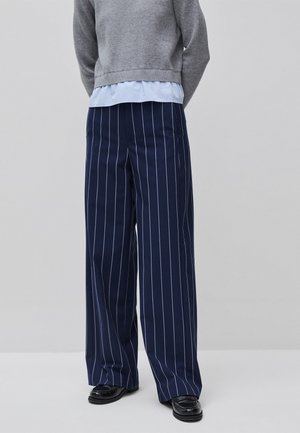 Marineblauwe broek met wijde pijpen en witte verticale krijtstrepen, gecombineerd met zwarte loafers, een grijze trui en een lichtblauw overhemd.