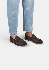 Mocasines de ante marrón con suela de goma plana y detalles de costura, combinados con jeans de denim azul claro.