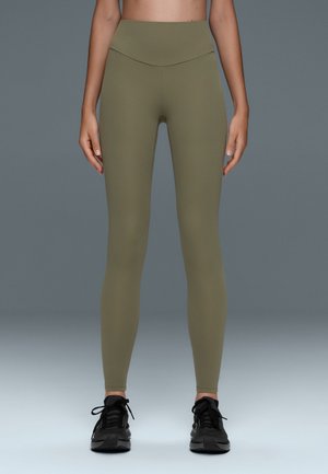 Osoba ubrana w oliwkowe legginsy z wysokim stanem i czarne buty sportowe stoi na szarym tle.