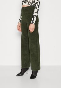 Groene corduroy broek met wijde pijpen en een zachte textuur, voorzien van achterzakken en een hoge taille, gecombineerd met zwarte puntige hakken.