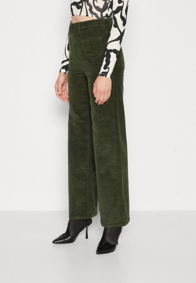 Groene corduroy broek met wijde pijpen en een zachte textuur, voorzien van achterzakken en een hoge taille, gecombineerd met zwarte puntige hakken.