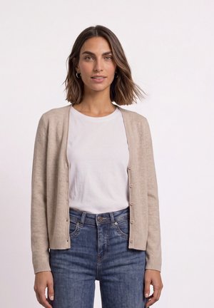 Femme aux cheveux bruns mi-longs portant un cardigan beige boutonné par-dessus un t-shirt blanc et un jean bleu sur un fond uni.
