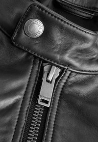 Nahaufnahme einer schwarzen Lederjacke mit einem silbernen Reißverschluss und einem Metallknopf, auf dem "SIGNATURE" eingraviert ist, und die glatte Textur sowie die Nahtdetails hervorhebt.