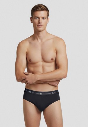 Giovane uomo con capelli corti castano chiaro, in piedi senza maglietta, indossa slip neri con fascia con marchio su sfondo uniforme.