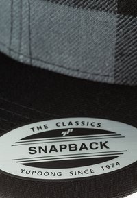 Snapback müts, millel on must kangas ja hall ruuduline muster. Lisatud on metalltag, millel on kiri "THE CLASSICS" ja "Yupoong Since 1974."