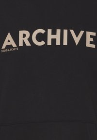 Sudadera negra con texto blanco en negrita: "ARCHIVE" en mayúsculas, con texto más pequeño "SOLID ARCHIVE" debajo en una fuente casual. Textura suave.