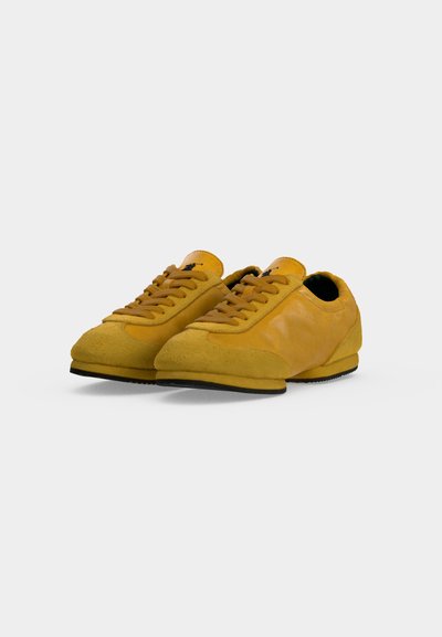 Paire de baskets jaunes en daim et cuir avec lacets et semelles en caoutchouc noires présentées sur un fond blanc uni.