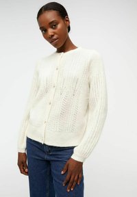 Gilet en tricot blanc avec un motif texturé, col rond et boutons en perles. Manches longues et coupe décontractée. Associé à un jean bleu foncé.