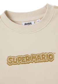 Sudadera beige con cuello redondo acanalado, con el logotipo en relieve "SUPER MARIO" en marrón y una textura suave. Etiqueta incluida en el cuello.