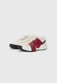 Nike-sko med en strukturert kremfarget overdel, røde detaljer og en svart gummisåle. Har en karakteristisk swoosh-logo i rødt.