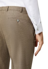 Hellbraune, strukturierte Hose mit einer Gesäßtasche und einer Knopfleiste, kombiniert mit einem weißen Langarmhemd, das über Manschettenknöpfe verfügt.