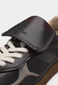 Σκούρο καφέ δερμάτινο αθλητικό παπούτσι με λευκά κορδόνια, ραμμένη φλοπ με την επιγραφή "ALOHAS" και λεπτομέρειες στη σόλα από σουέντ και καουτσούκ.