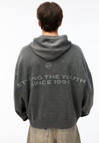 Personne portant un hoodie noir délavé avec la capuche abaissée, texte "STYLE LA JEUNESSE DEPUIS 1991" dans le dos, et un pantalon beige clair.