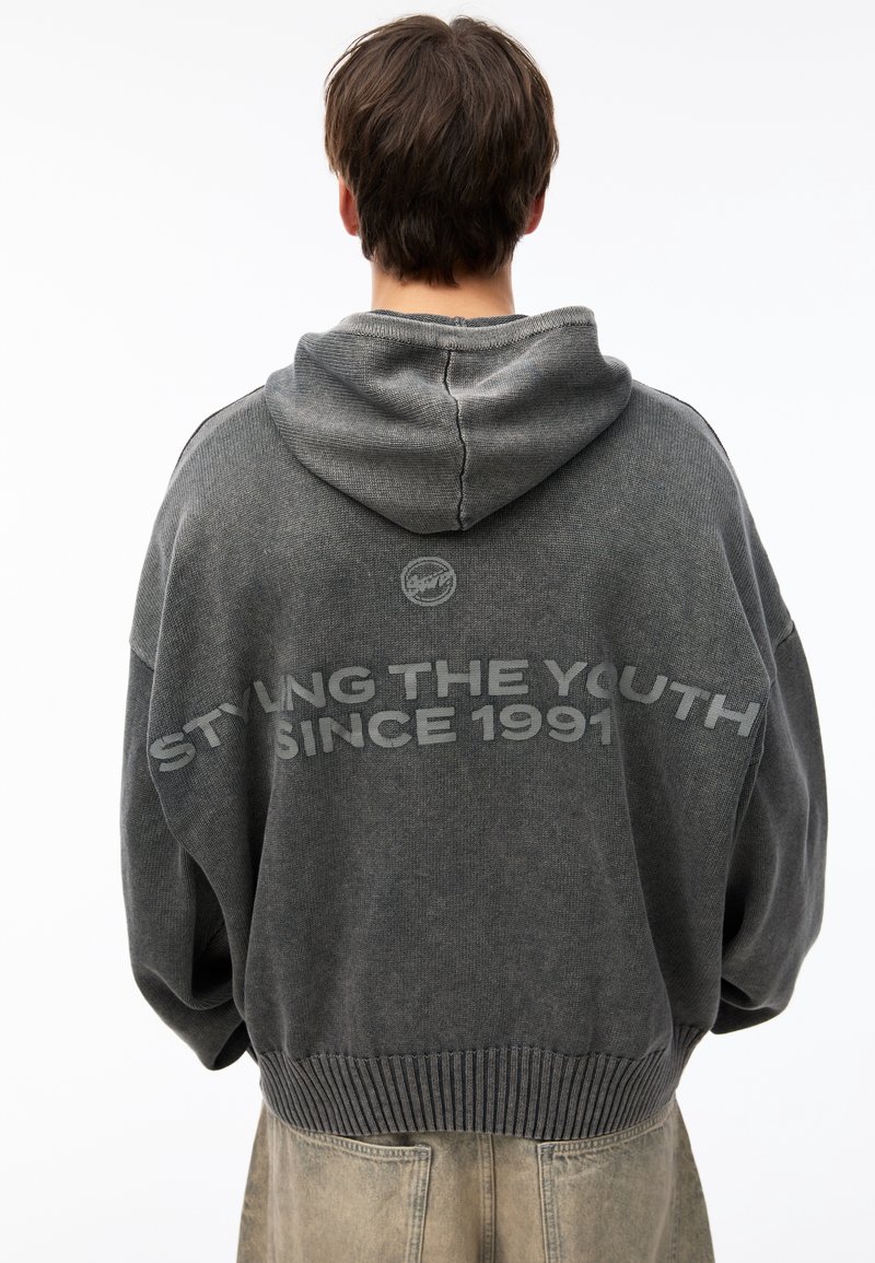 Personne portant un hoodie noir délavé avec la capuche abaissée, texte "STYLE LA JEUNESSE DEPUIS 1991" dans le dos, et un pantalon beige clair.
