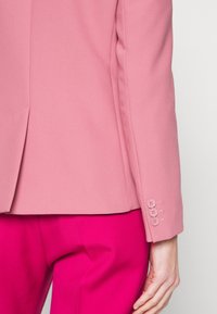 Blazer rosa ajustado con un tejido suave, que presenta un detalle de puño con tres botones. Llevado sobre pantalones rosa brillante.