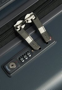 Maleta rígida gris con cremallera de tiradores metálicos dobles, panel de candado de combinación y superficie texturizada. Incluye un orificio para llave para mayor seguridad.