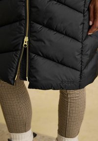 Czarna puchowa kurtka z przeszyciami w kształcie litery V i złotym suwakiem. Marka "Joules" jest wyszyta na czarno na dolnym brzegu. Pod spodem legginsy w wzór houndstooth.