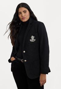Črn blazer s srebrnimi gumbi in vezenim logotipom na žepu. Ima prilegajoč kroj in ovratnik s zarezo. Obučen je nad črnim jopičem.