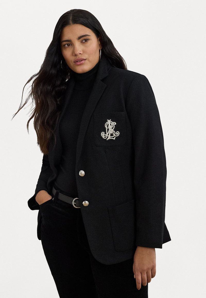 Črn blazer s srebrnimi gumbi in vezenim logotipom na žepu. Ima prilegajoč kroj in ovratnik s zarezo. Obučen je nad črnim jopičem.