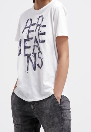 Person iført hvid T-shirt med dristige sorte abstrakte bogstaver og mørkegrå vaskede jeans, hænder i lommerne, foran en ensfarvet baggrund.