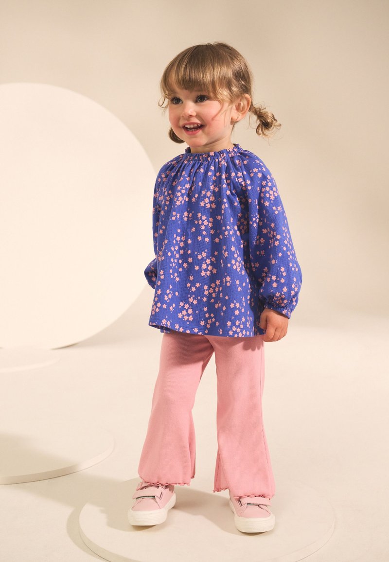 Blusa azul con estampado floral y mangas abullonadas, combinada con pantalones acampanados de canalé rosa y zapatillas de color claro sobre un fondo neutro.