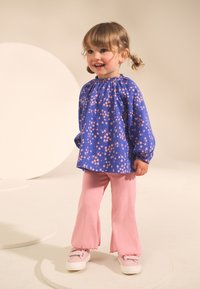 Blusa blu con motivo floreale e maniche a sbuffo, indossata con pantaloni svasati rosa a coste e sneakers di colore chiaro su uno sfondo neutro.