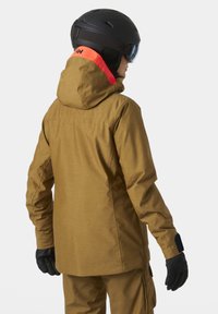 Chaqueta de esquí impermeable marrón con capucha, que presenta un collar de color naranja brillante, guantes negros y un casco de esquí, mostrada desde un ángulo trasero.