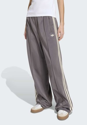 Persona che indossa pantaloni della tuta Adidas grigi oversize con strisce bianche e logo, abbinati a scarpe da ginnastica bianche e una maglietta bianca.