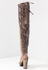 Botte haute à talons, dépassant le genou, avec un motif serpent marron et noir et une fermeture à lacets en haut, sur fond uni.