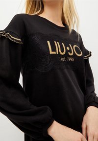Svart sweatshirt med guldtexten "LIU·JO est. 1995," spetsdetalj fram och volangärmar. Mjuk textur, avslappnad passform.