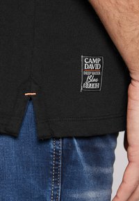 Czarna bawełniana koszulka z etykietą z białym i pomarańczowym tekstem, detalem o wydzielonym bocznym szwie oraz akcentami w szwie, w połączeniu z niebieskimi dżinsami.