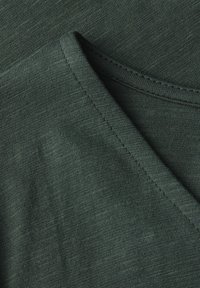 Tmavozelené tričko s výstřihem do V, vyrobené z jemného materiálu. Límec a spodní lem mají čisté švy, což podtrhuje ležérní design.