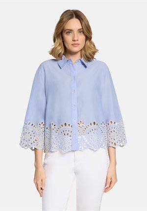 Frau trägt eine hellblaue, geknöpfte Bluse mit wellenförmigen floralen Spitzenbesatz am Saum und weiten Ärmeln, kombiniert mit weißen Hosen.