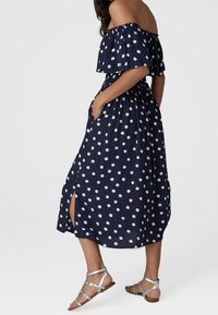 Žena oblečená v námořnických midi šatech s ramínky, pokrytá puntíky, s bočními rozparky a stříbrnými plochými sandály se řemínky, má ruce v kapsách a stojí na bílém pozadí.