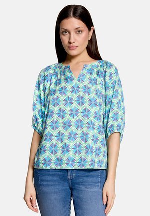 Femme portant un chemisier à motif géométrique multicolore avec des manches mi-longues et une encolure fendue, associé à un jean bleu.