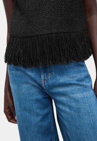 Top negro texturizado con detalles de flecos en el dobladillo, combinado con vaqueros de denim azul claro. Costuras visibles y un ajuste casual.