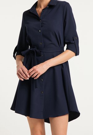 Žena nosící tmavě modré košilové šaty na knoflíky se tříčtvrtečními rukávy a zavázaným páskem v pase, stojící s lehce sepjatýma rukama.